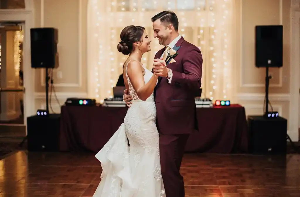 Bruidspaar tijdens eerste dans op verlichte dansvloer, met bokeh van lichtjes en dj-opstelling op elegante trouwlocatie.