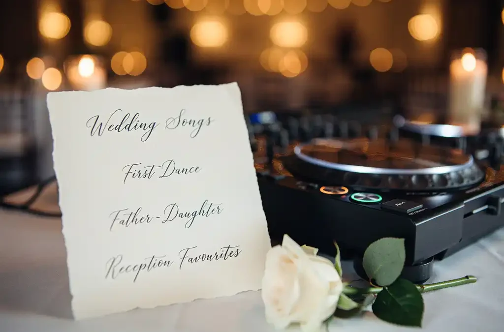 Handgeschreven bruiloft songslijst op crèmepapier naast een DJ-controller en witte roos, met warme kaarslichtsfeer op de achtergrond.